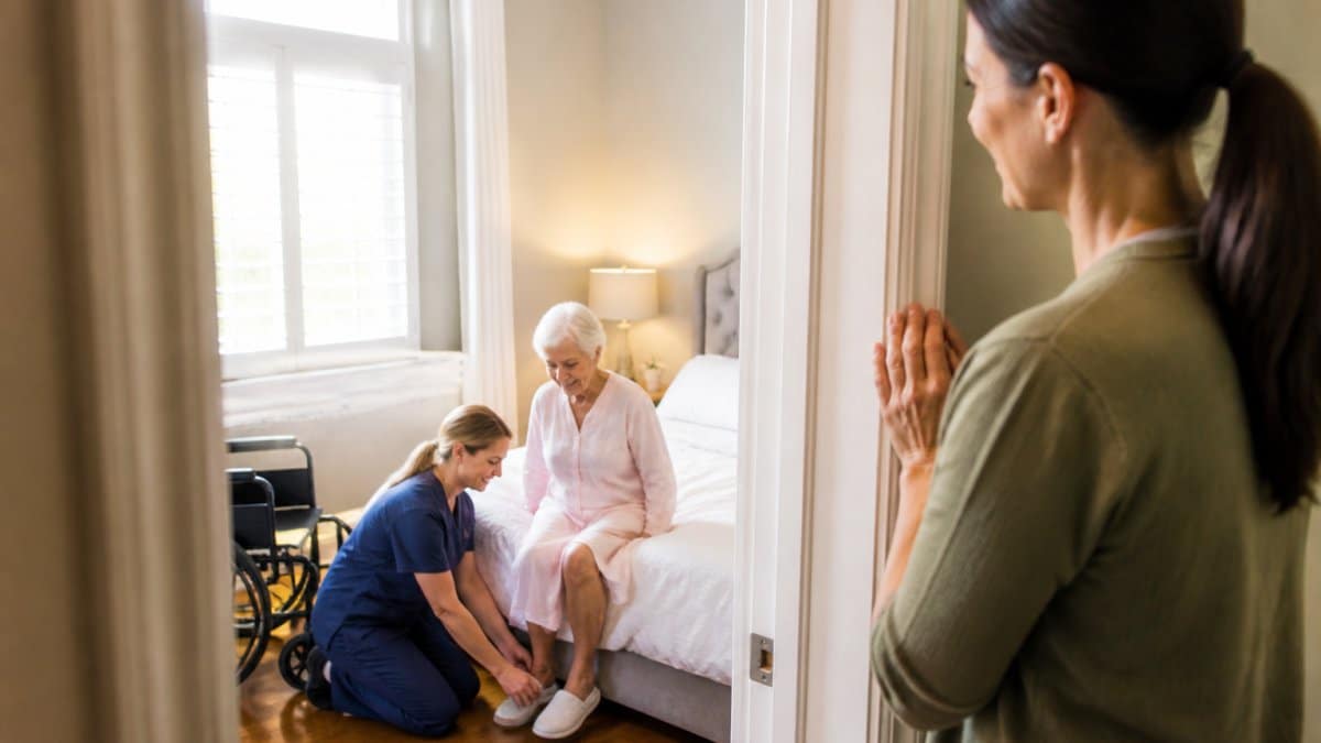 Professional caregiver assisting an older adult with daily activities while a family member watches from the doorway, appearing relieved and grateful, in a bright home setting
